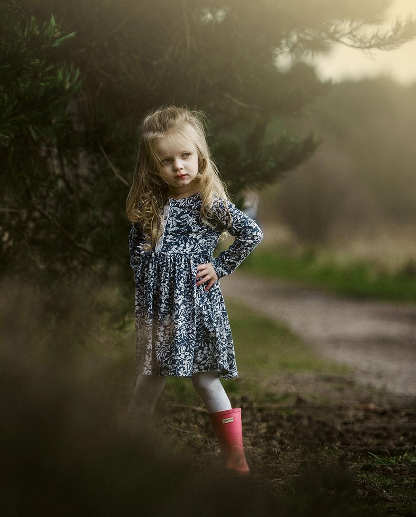 A young girl with long blonde hair stands outdoors on a dirt path, wearing a patterned dress, white leggings, and bright pink rain boots. She poses confidently with one hand on her hip near tree branches.