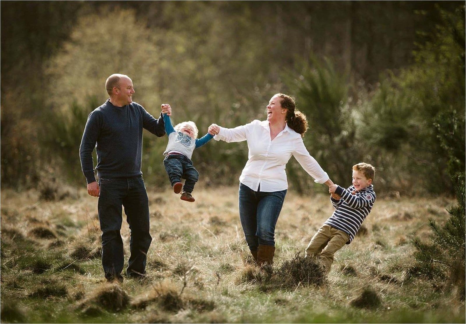 A smiling family of four plays outdoors; two parents swing a young child between them while an older child holds the mothers hand, all standing on grassy terrain surrounded by trees.