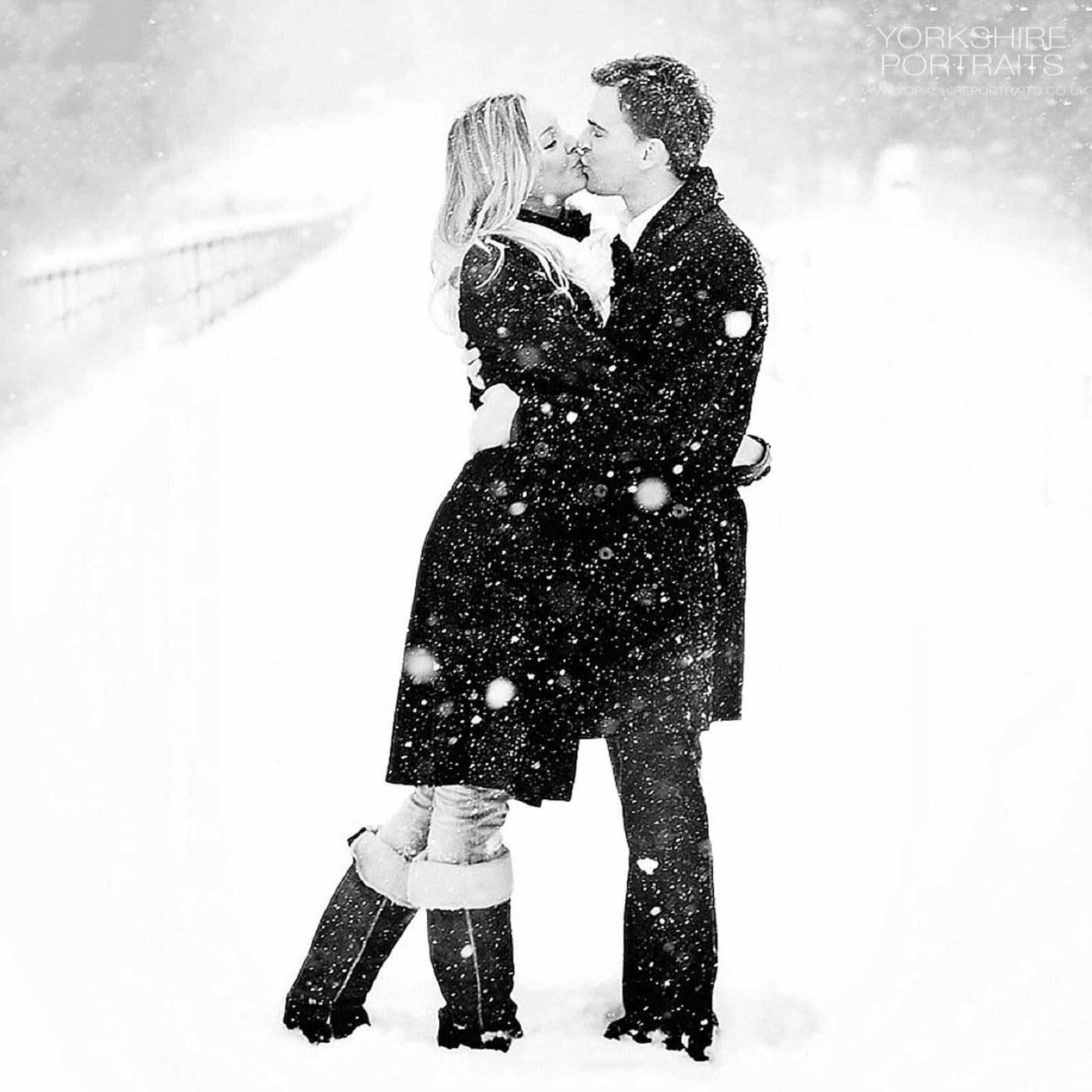 A couple dressed in winter coats and boots kisses in the snow, surrounded by falling snowflakes on a snowy path. The scene is romantic and wintry, with a soft, white background.