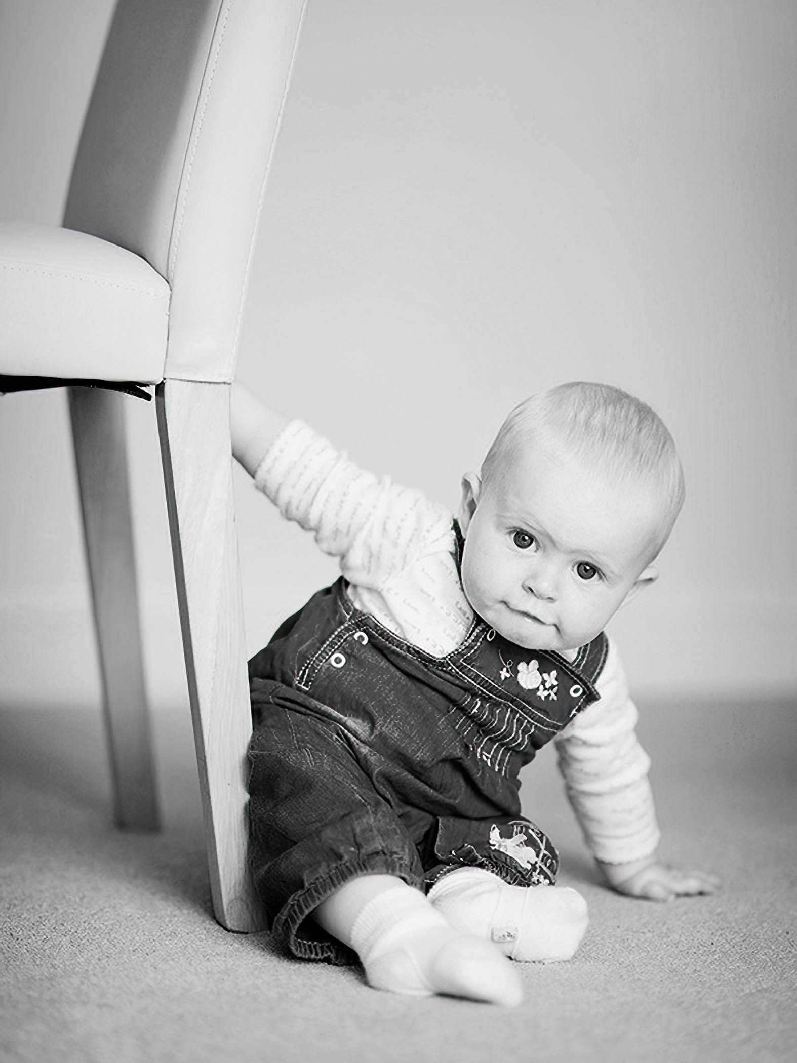 A baby wearing denim overalls and socks sits on the floor, holding onto the leg of a chair and looking curiously at the camera. The image is in black and white.
