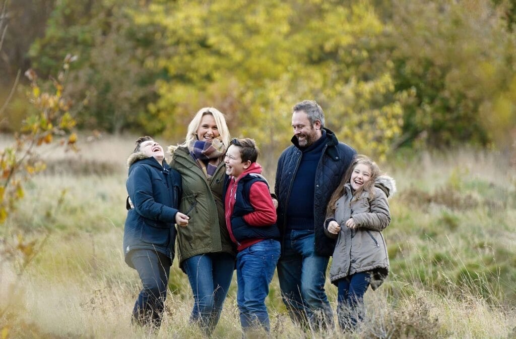 A family of five, dressed in jackets, stands together laughing in a grassy field with autumn trees in the background. They appear happy and are enjoying time outdoors.