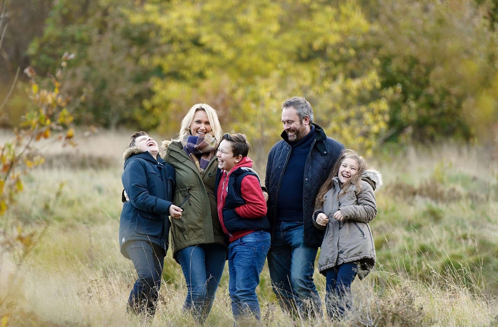 A family of five, dressed in jackets, stands together laughing in a grassy field with autumn trees in the background. They appear happy and are enjoying time outdoors.