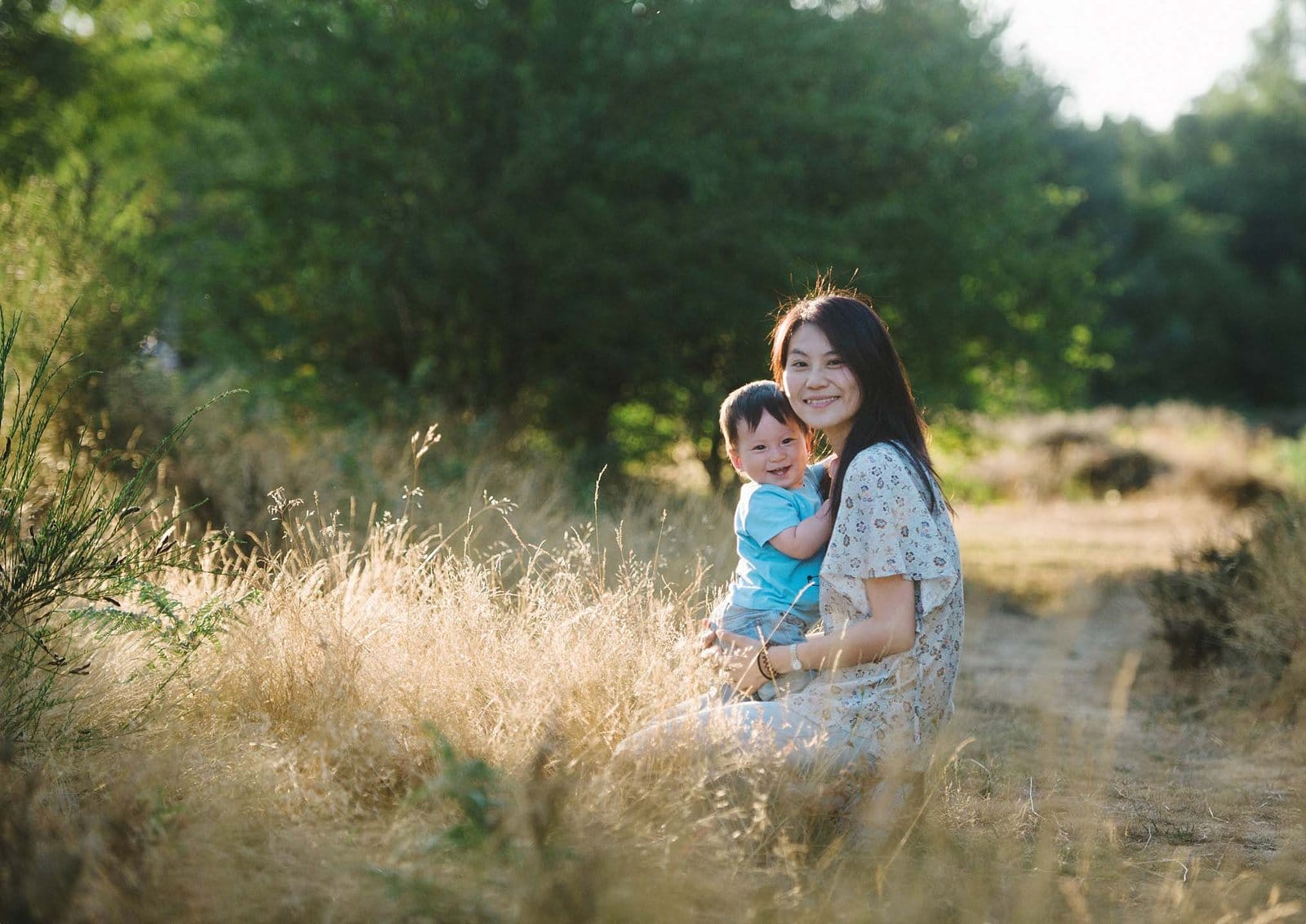 A woman in a light floral dress sits in a sunny, grassy field holding a smiling baby. Both are looking at the camera, surrounded by green trees and golden sunlight.