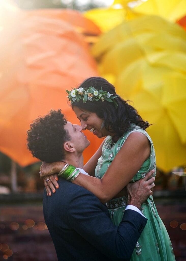 A man in a suit lifts and embraces a smiling woman in a green dress and floral crown. They gaze joyfully at each other, with large yellow and orange decorations blurred in the background.