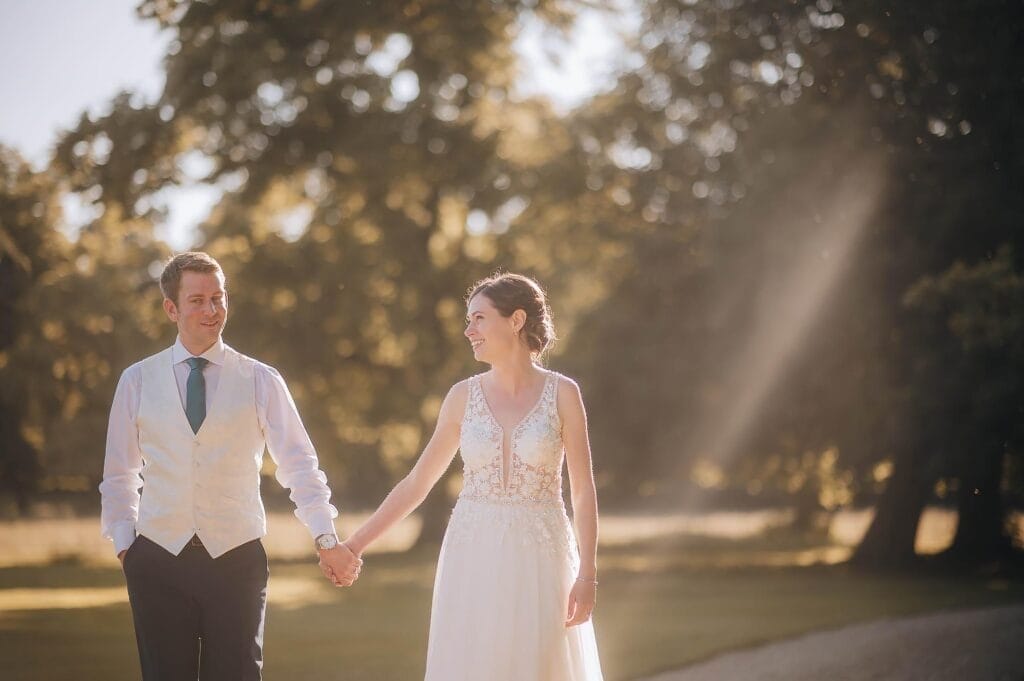 A bride and groom hold hands and smile at each other while walking outdoors on a sunny day, surrounded by trees and soft sunlight.