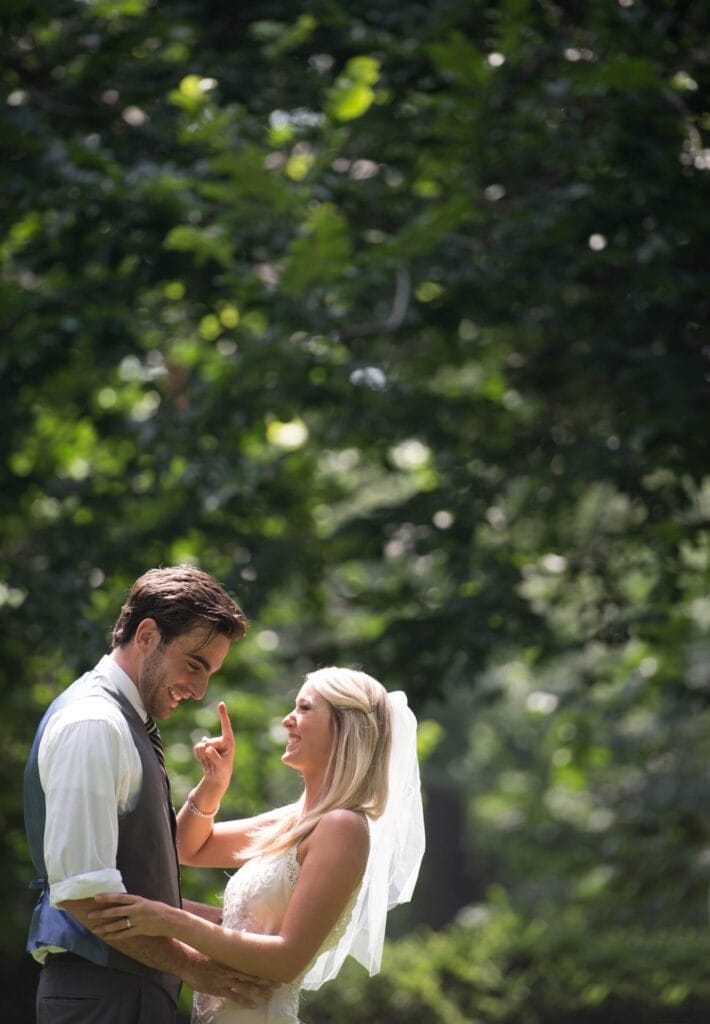 A bride and groom smile at each other under a canopy of trees. The bride wears a white dress and veil, and the groom is in a white shirt and gray vest. Sunlight filters through the leaves, illuminating the couple—a perfect moment captured by a talented Doncaster wedding photographer.
