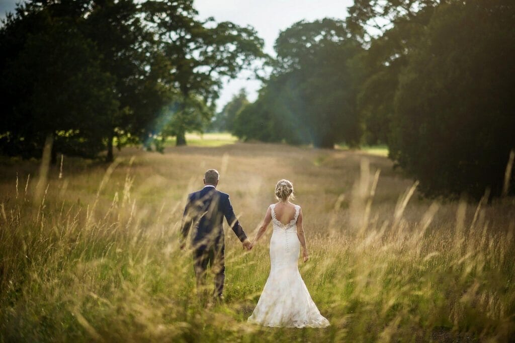 A bride and groom walk hand in hand through a sunlit grassy field, surrounded by tall trees and golden grass, captured from behind on their wedding day.