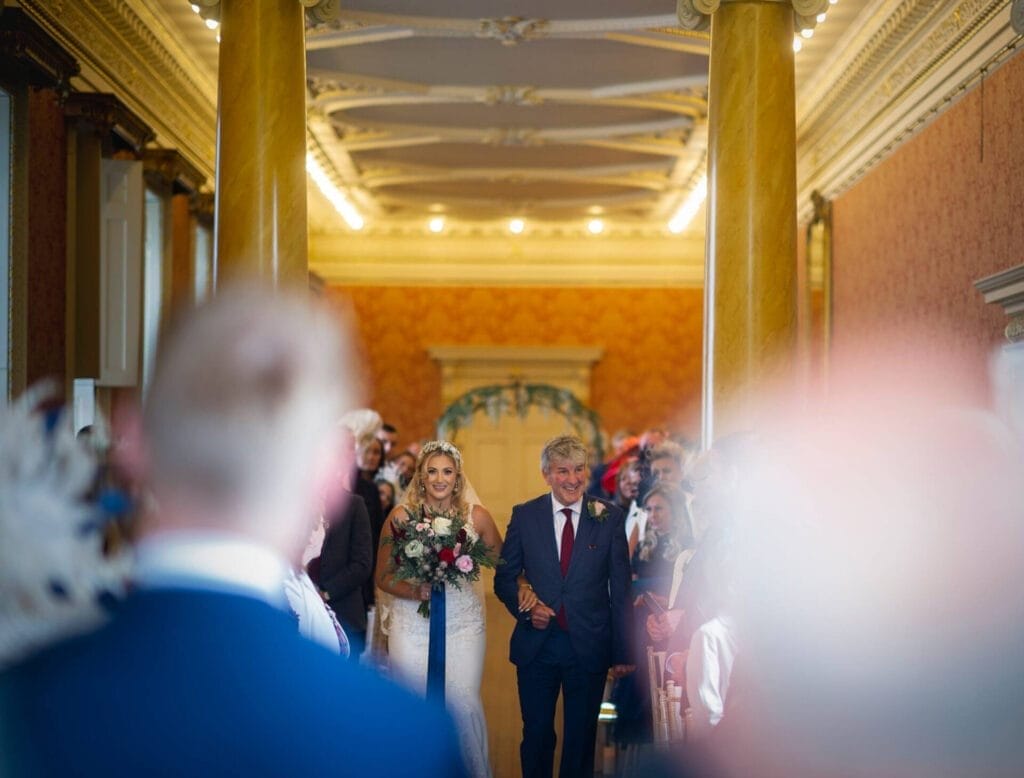 A bride in a white dress holding a bouquet walks down the aisle with an older man in a suit. Guests watch from both sides, and warm lighting fills the ornate hall. The foreground is blurred.