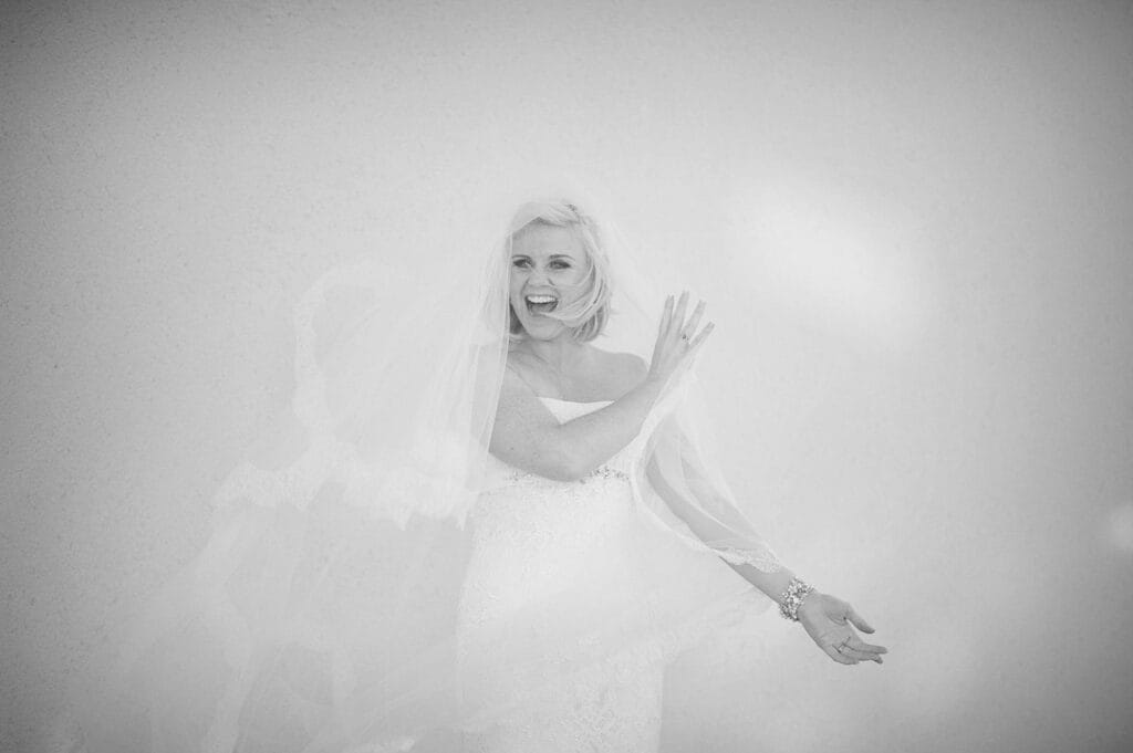 A joyful bride in a strapless wedding dress and veil smiles and laughs, holding her veil with one hand against a plain, light background. The image is in black and white.