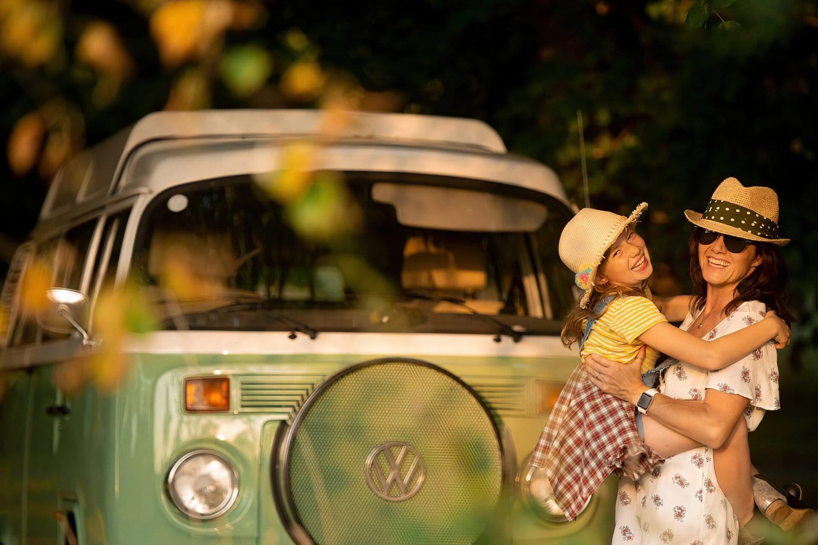 A woman and a young girl wearing hats smile and embrace in front of a vintage green Volkswagen van, surrounded by greenery and warm sunlight.