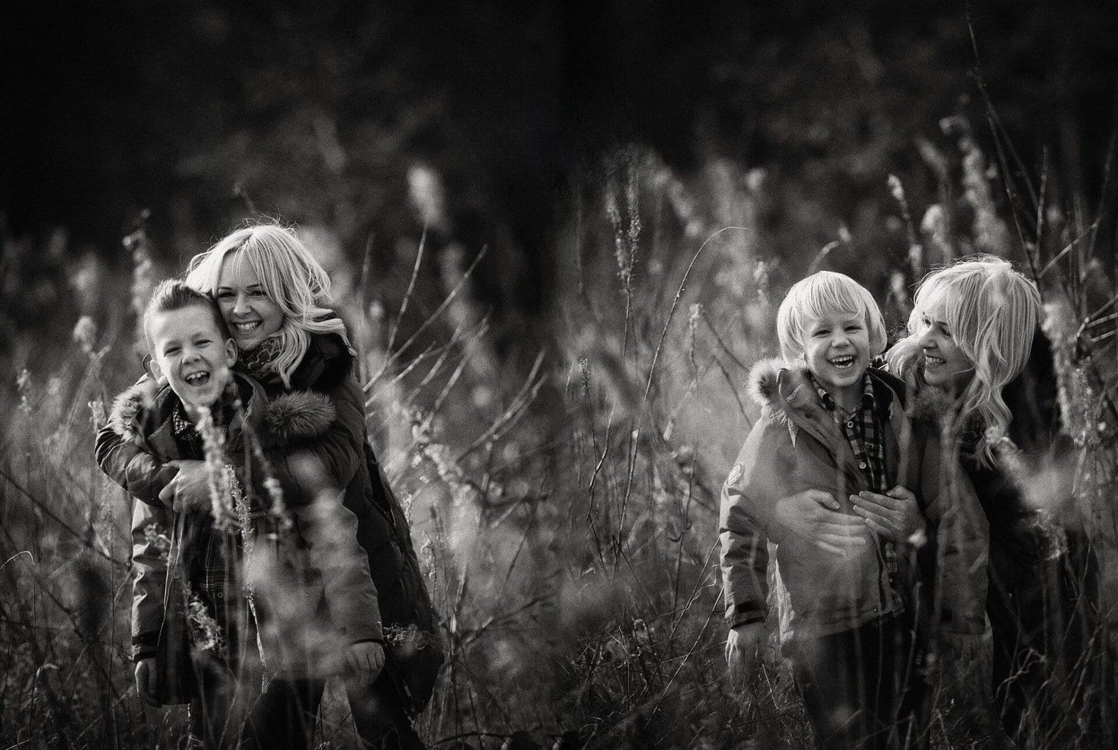 A black-and-white photo shows a woman with long blonde hair laughing and playing with two young boys in a field of tall grass. Everyone is wearing coats, and the mood is joyful and carefree.