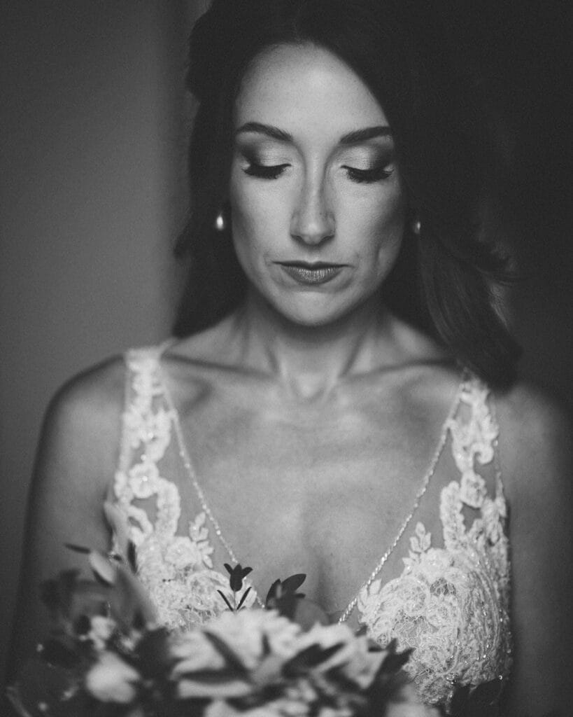 Black and white photo of a woman in a lace dress, looking down thoughtfully while holding a bouquet. The focus is on her face and upper body, creating a soft, intimate atmosphere.