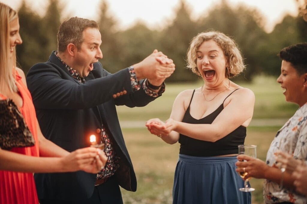 A man dazzles three surprised onlookers with a magic trick outdoors. As he seemingly makes an object vanish in his cupped hands, one person holds a lit candle and another a glass, creating a scene as enchanting as those captured by a Yorkshire Wedding Photographer. Lush trees and grass complete the setting.
