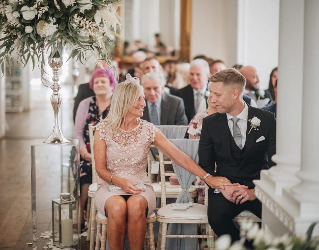 A woman in a light pink dress sits smiling and holding hands with a man in a suit at a wedding ceremony, surrounded by seated guests and elegant floral decorations.