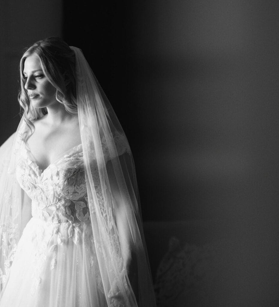 A bride in a lace wedding gown and veil stands in soft light, looking to the side with a serene expression. The black-and-white photo captures her in a moment of quiet reflection.