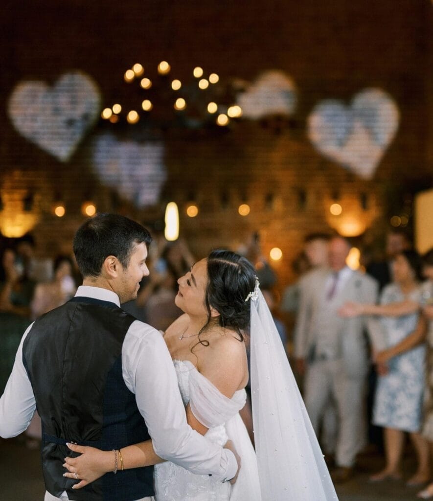 A bride and groom share a dance, smiling at each other, with guests watching in the background. Soft lighting and large heart-shaped decorations are visible on the brick wall behind them.