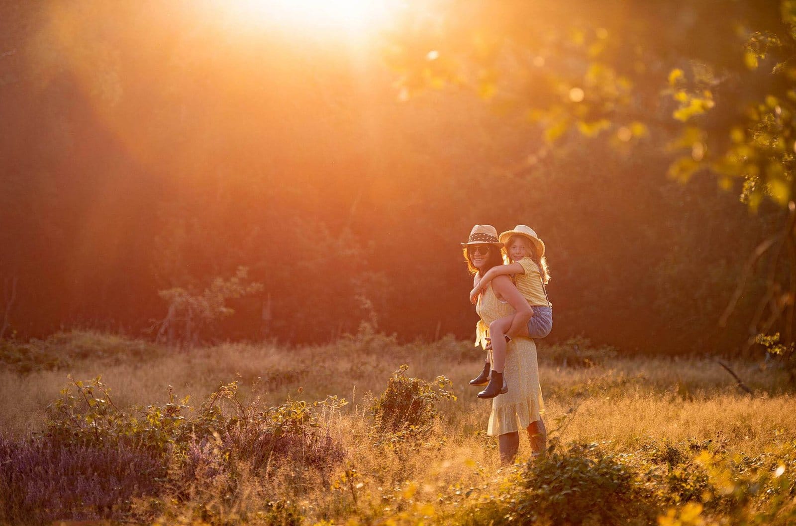 A woman in a hat gives a piggyback ride to a smiling girl in a sunlit meadow, surrounded by tall grass and golden light in the late afternoon.