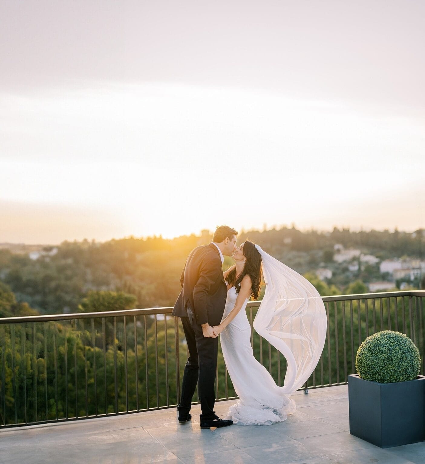 Yorkshire Wedding Photographer - Yorkshire Portraits 26 A couple kisses on a balcony during sunset at their Florence destination wedding. The brides long veil flows in the breeze as the groom stands in a black suit. Nearby, a small potted shrub complements the scenic view of trees and hills—a perfect capture by a Villa Cora wedding photographer.