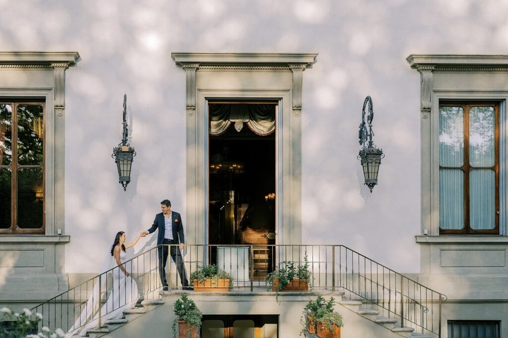 A bride and groom stand on an elegant staircase outside a large, ornate building. The groom holds the brides hand as she descends the stairs. Captured beautifully by a UK destination wedding photographer, the scene is framed by large windows and decorative lanterns, with potted plants lining the steps.