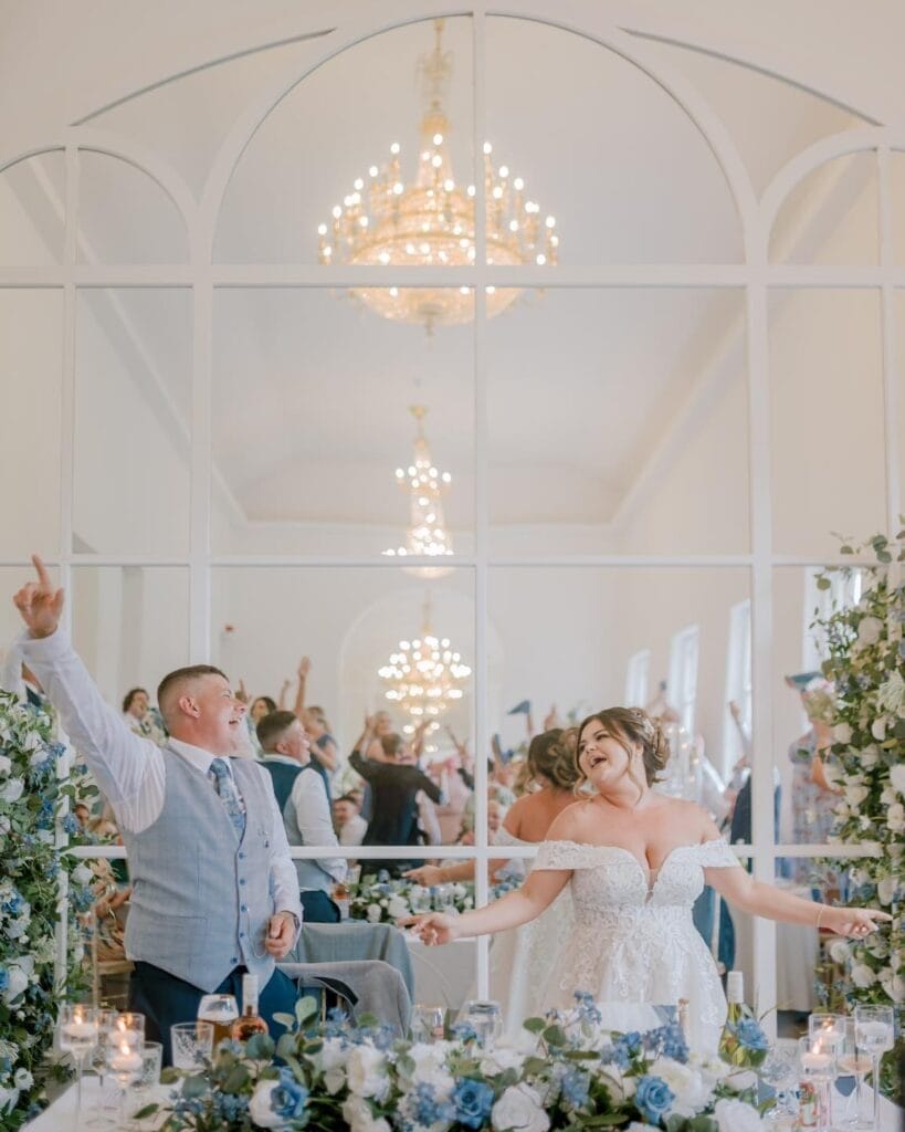 A bride and groom joyfully raise their arms during a wedding reception captured by a Norwood Park wedding photographer. They are surrounded by floral arrangements with blue and white flowers, and a large chandelier is reflected in the mirrored wall behind them.