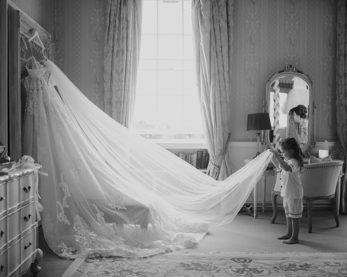 In a well-lit room bathed in elegance, a little girl gently holds the long train of a wedding dress draped over a wardrobe, admiring its beauty. Captured in black and white by a Norwood Park wedding photographer, the scene features elegant decor including a large window, mirror, and chair.