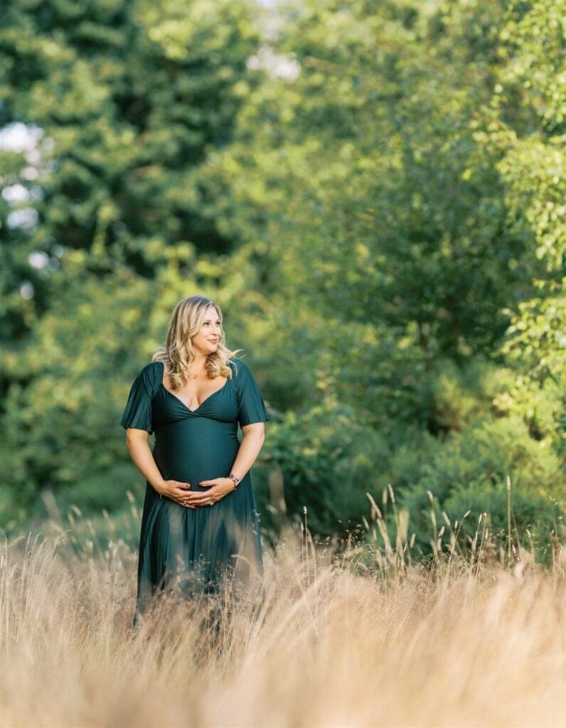 A pregnant woman in a dark green dress stands in tall grass, cradling her belly and smiling, surrounded by lush green trees in a sunlit outdoor setting.