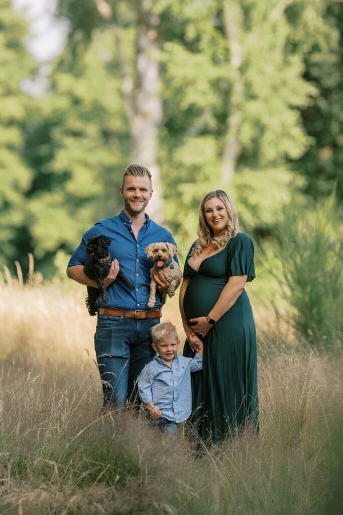 A smiling family stands outdoors in tall grass. The man holds two small dogs, the woman in a green dress rests her hand on her pregnant belly, and a young boy stands in front of them. Trees and greenery fill the background.