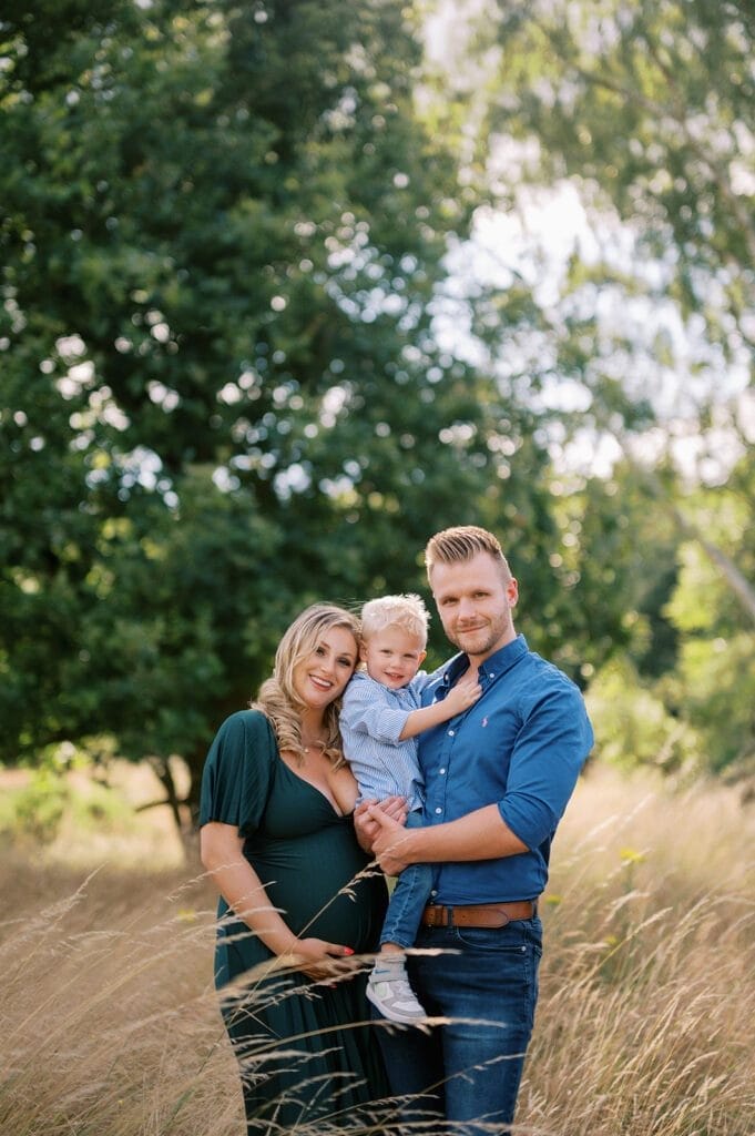 A smiling pregnant woman in a green dress stands next to a man in a blue shirt holding a young child. They are outdoors in a grassy field with tall trees in the background, all looking at the camera.