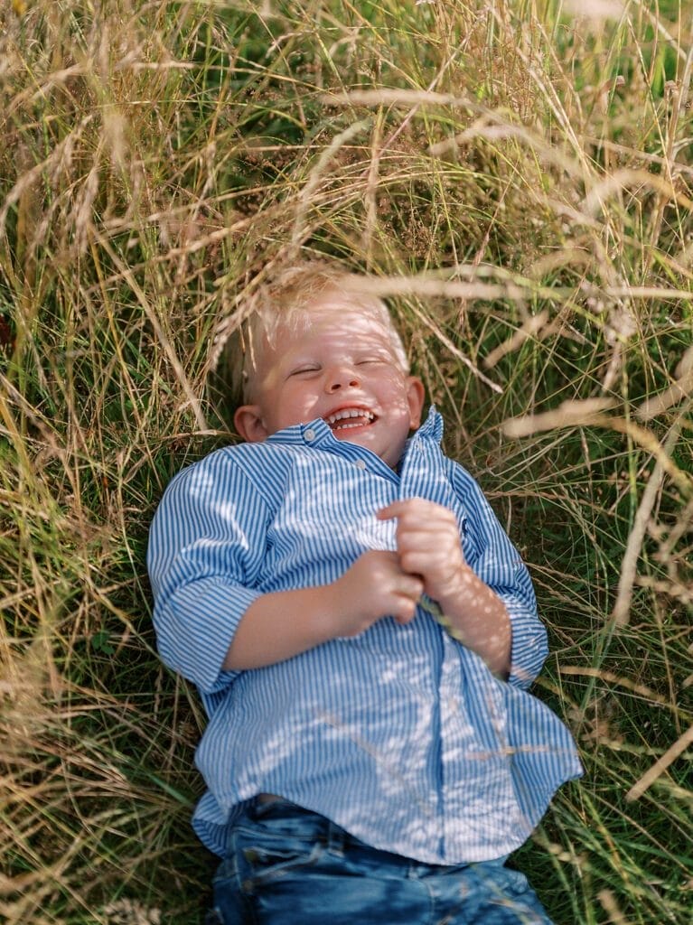 A young boy wearing a blue striped shirt and jeans lies in tall grass, smiling and laughing during a pregnancy family photoshoot at Clumber Park, with sunlight filtering through the blades around him.