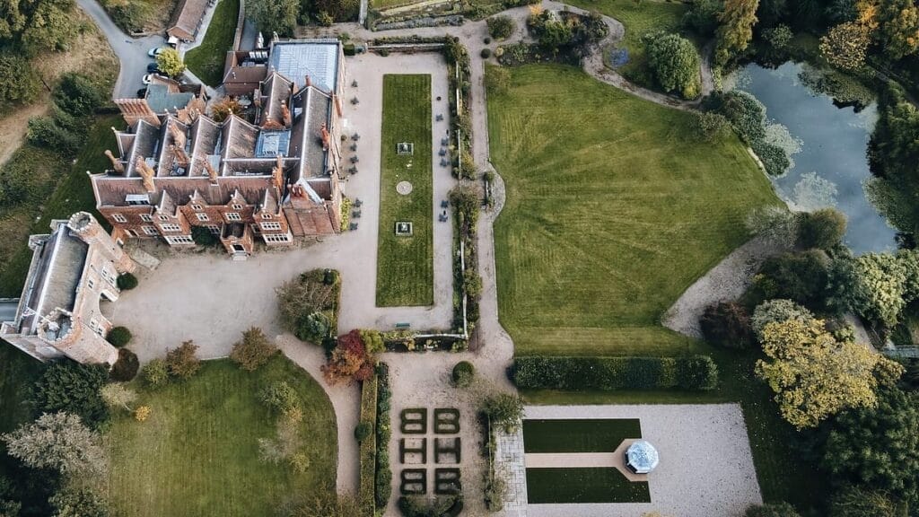 Aerial view of an estate with a large brick mansion surrounded by landscaped gardens, perfect for Nottinghamshire wedding photography. The grounds include neatly trimmed lawns, pathways, hedges, and a small pond. The gardens feature geometric patterns and lush greenery.