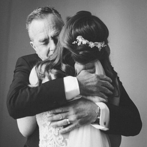 A black-and-white photo captures the essence of Yorkshire wedding photography, with a bride and an older man hugging tightly. The brides back reveals her lace dress and floral hairpiece, while the mans face is visible, eyes closed, conveying a tender expression.