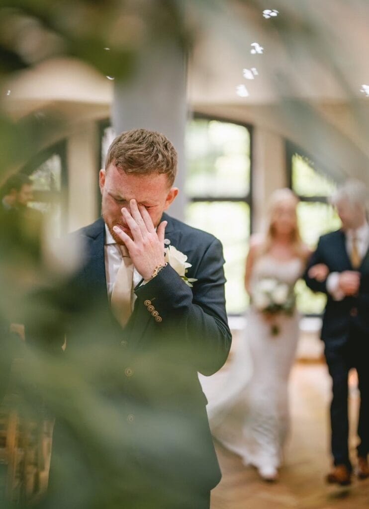 A man in a suit wipes his eye at the Ollerton Pumping House, with blurred greenery in the foreground. In the background, a woman in a wedding dress walks down the aisle with an older man by her side, holding a bouquet. The scene captured by an Ollerton wedding photographer is emotional and intimate.