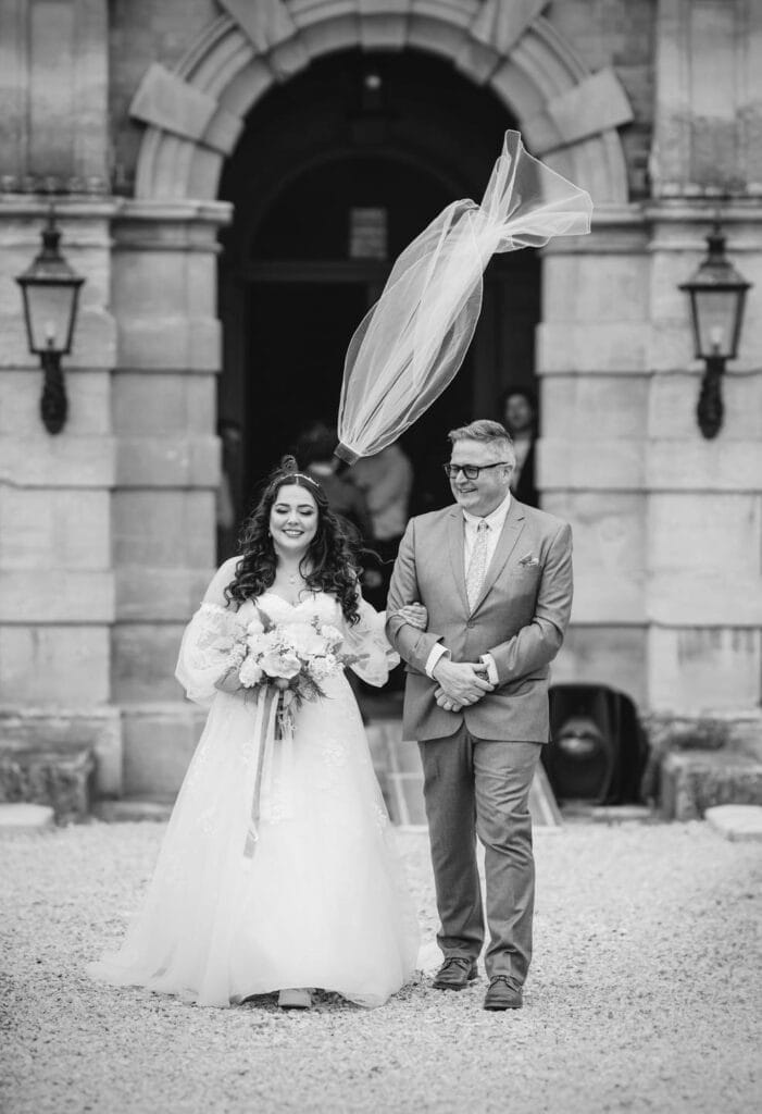Yorkshire Wedding Photographer - Yorkshire Portraits 11 A bride, bouquet in hand, walks withher father in a suit past a stone building, her veil caught mid-air. Captured by a Rossington Hall wedding photographer, this enchanting scene comes alive in timeless black and white.