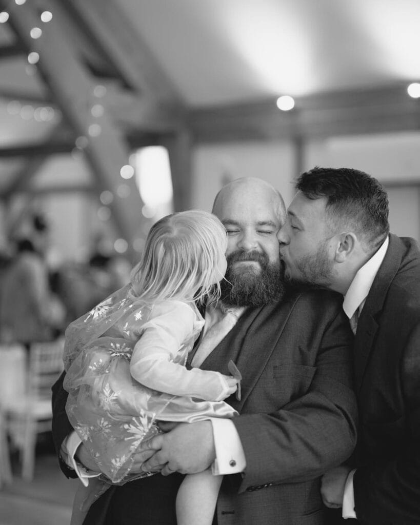 In a warmly lit indoor setting, a bearded man smiles while holding a young child in a dress with a sheer embroidered overlay. Another man in a suit affectionately kisses the bearded mans cheek, capturing the joy of the moment perfectly, as seen through North Yorkshire wedding photography.