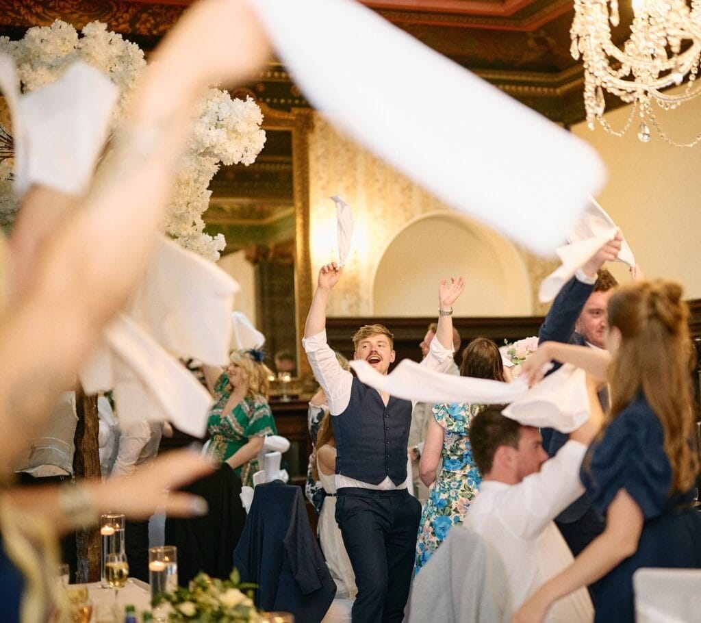 A joyful man in a blue vest enthusiastically waves a napkin amidst lively wedding guests at Wortley Hall. Others join in under the ornate chandelier, celebrating with gusto in the elegantly decorated venue, perfectly captured by the talented wedding photographer.
