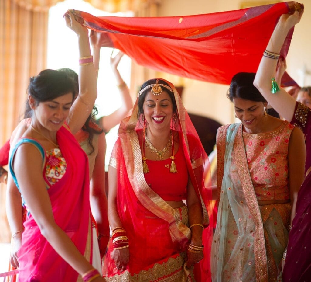 A joyful bride in a red saree smiles as she walks under a red cloth held by women in colorful sarees. They appear to be participating in a traditional wedding ceremony, surrounded by warm light and festive décor.