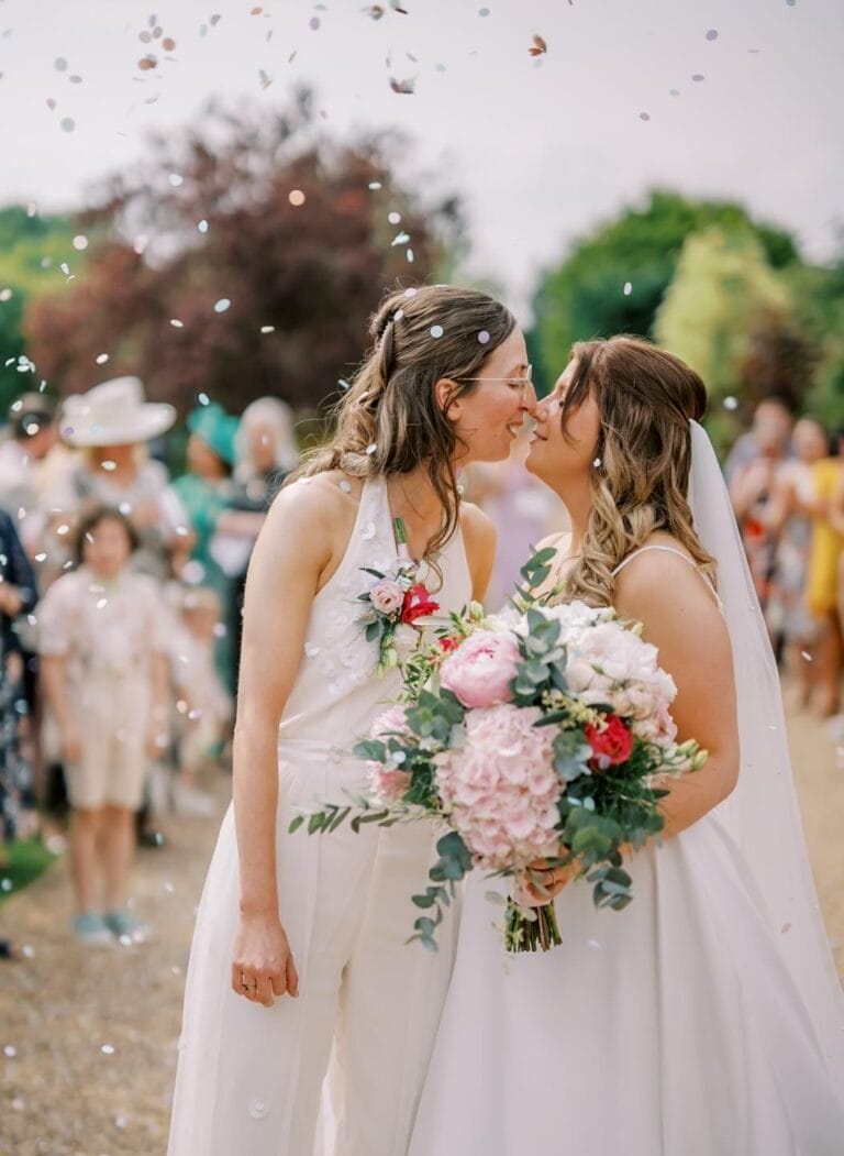 Two brides in wedding attire share a kiss outdoors at a picturesque Nottinghamshire wedding venue while guests throw confetti. One bride holds a lush bouquet of pink and white flowers, against a backdrop of blurred greenery and smiling guests celebrating their love.