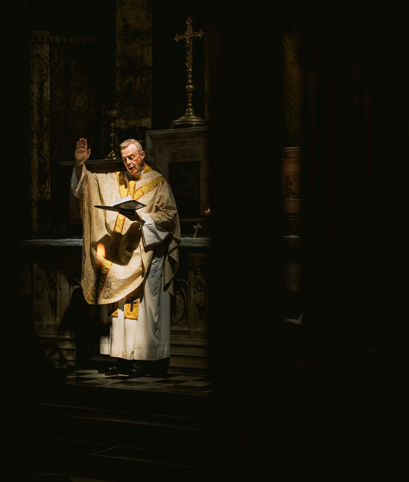 A priest in ornate robes stands near an altar, raising one hand in blessing while holding a book in the other. Soft, dramatic lighting highlights him against a dark background, with a cross visible behind.