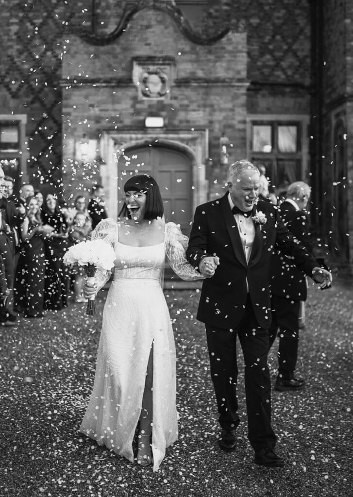 Black and white photo of a joyful couple in formal attire walking outside a building. The woman wears a gown and holds flowers, and the man is in a suit. They smile as confetti falls around them, with guests celebrating in the background.