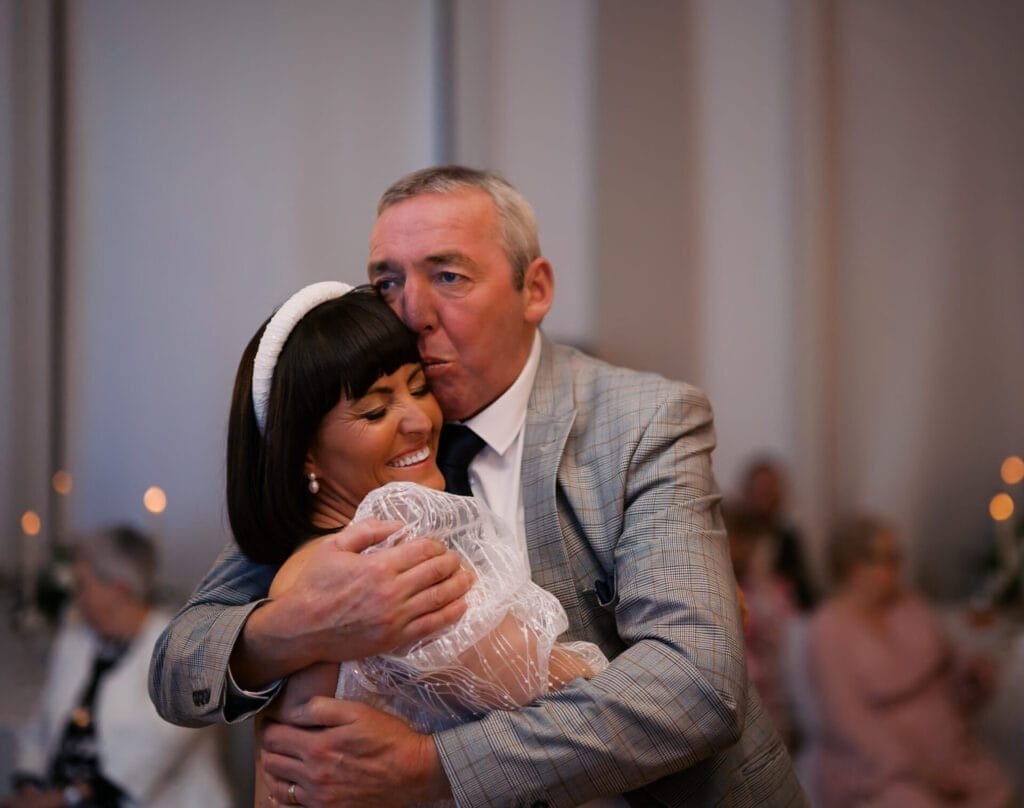 A man in a suit affectionately hugs a woman in a white dress, kissing her on the forehead. The background features soft lighting and blurred figures seated at tables, suggesting a celebratory event. Both appear joyful.