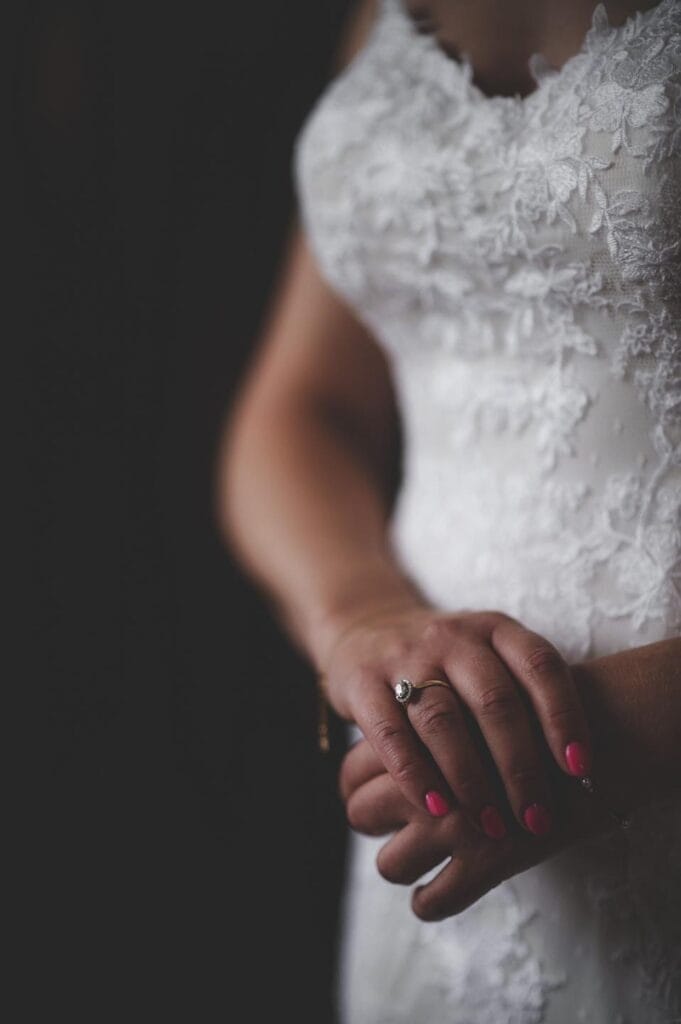 A person wearing a white lace wedding dress is seen from the shoulders down, hands clasped in front, showcasing pink nails and a ring on their left hand. The dramatic contrast against the dark background captures the essence of Rossington Hall wedding photography.