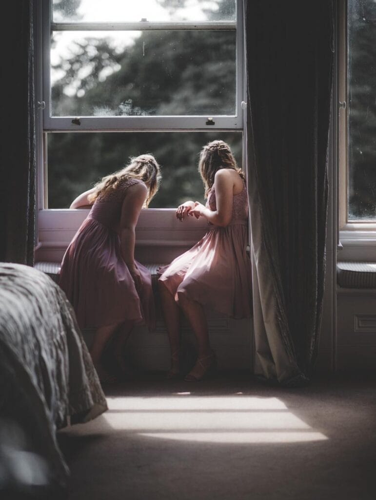 Two women in pastel dresses lean out of a large window, gazing outside. One is seated on the windowsill, while the other bends forward. Soft light from outside bathes the room, creating a serene atmosphere.