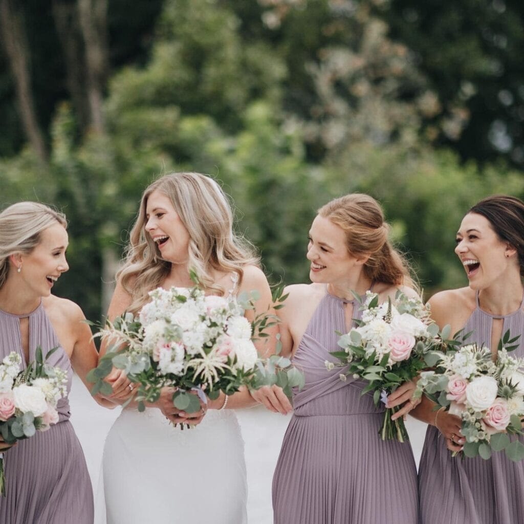 A bride in a white dress stands outdoors with three bridesmaids in lavender dresses, all laughing and holding bouquets of pink and white flowers. Green foliage provides a picturesque backdrop, perfectly captured by a Yorkshire wedding photographer.