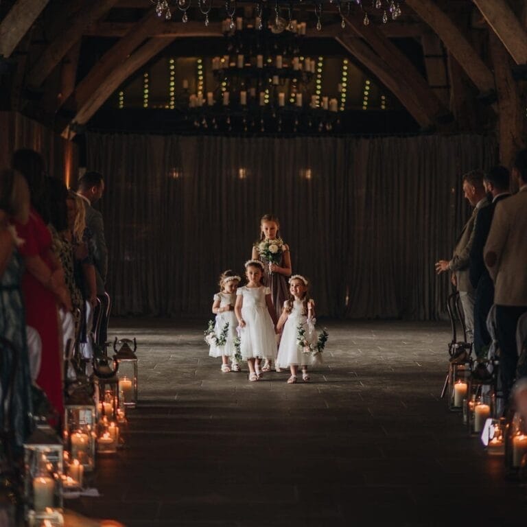 Four young flower girls in white dresses walk down an aisle lit by candles inside a rustic venue with wooden beams. Captured by a Yorkshire wedding photographer, guests watch as the leading girl, holding a bouquet, leads others adorned with flower crowns.