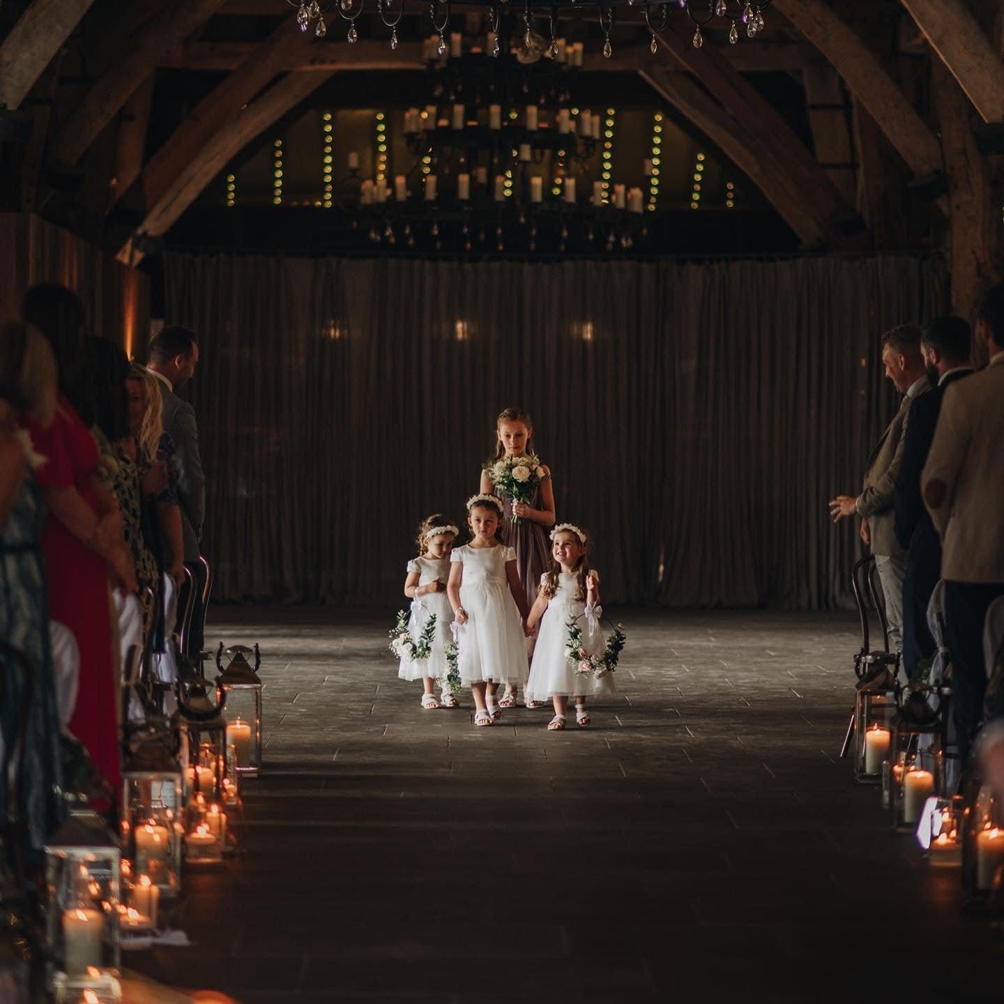 Four young flower girls in white dresses walk down an aisle lit by candles inside a rustic venue with wooden beams. Captured by a Yorkshire wedding photographer, guests watch as the leading girl, holding a bouquet, leads others adorned with flower crowns.