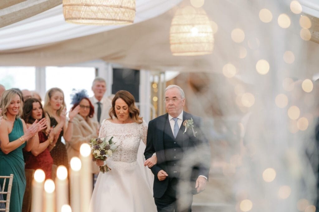 At the Woodman Inn, a bride in a white gown gracefully walks down the aisle with an older man in a suit. Guests stand and clap as the wedding photographer captures the moment. Soft lighting and decorative elements create a warm atmosphere at this Thunderbridge venue.