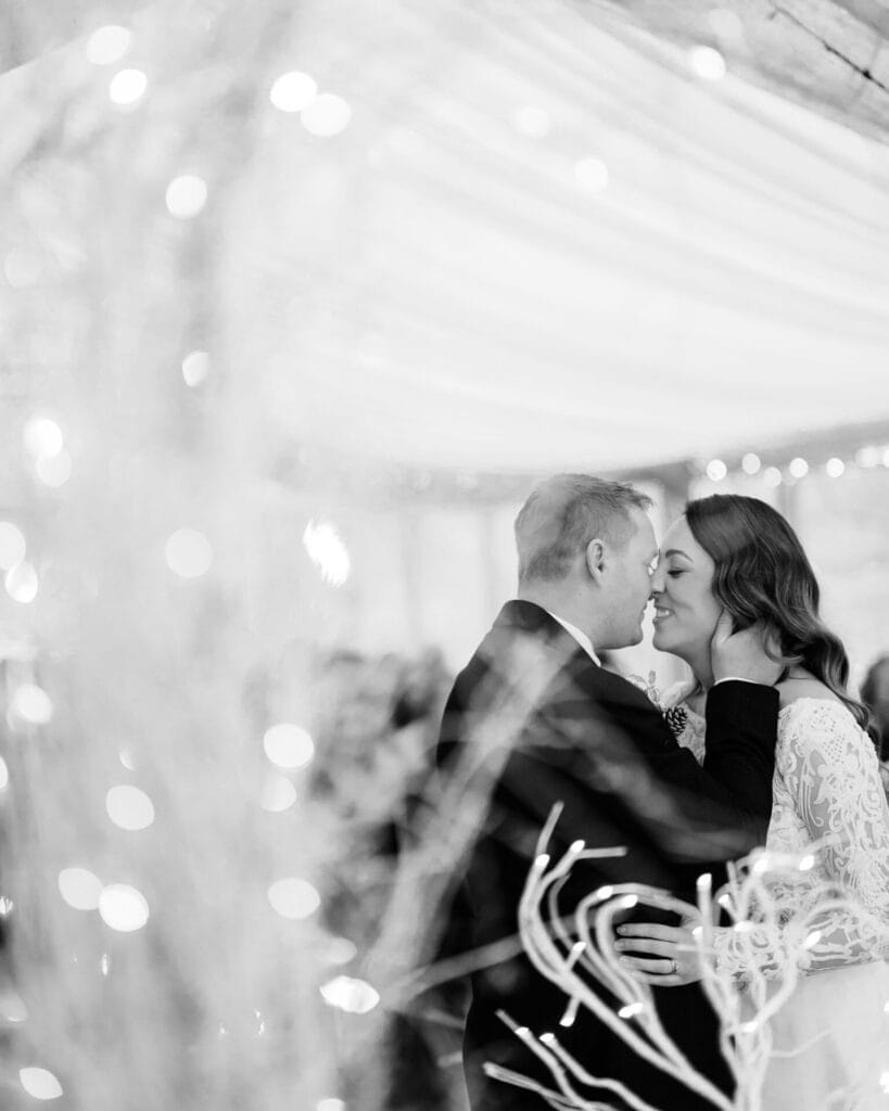 A black and white photo captures a couples intimate moment at their Thunderbridge wedding. The groom, in a suit, embraces the bride in her lace dress. Soft drapes and blurred lights enhance the romantic atmosphere, skillfully immortalized by The Woodman Inn wedding photographer.