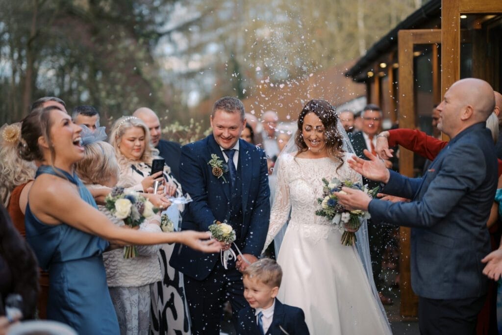 A bride and groom, smiling broadly, walk through a crowd at The Woodman Inn. Guests toss confetti into the air. The bride holds a bouquet as a young child stands near. Captured by a wedding photographer at Thunderbridge, everyone appears joyous amidst greenery and charming buildings.