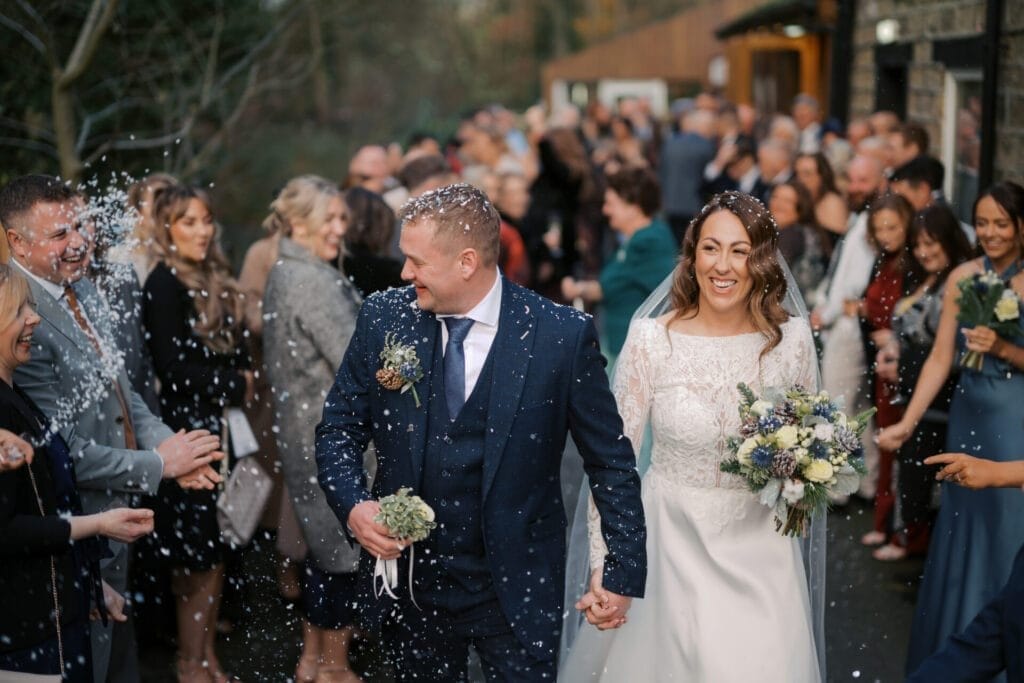 A bride and groom walk hand in hand through a crowd of smiling guests at Thunderbridge, who are tossing confetti. The bride holds a bouquet of flowers and wears a white dress, while the groom sports a blue suit. Captured by The Woodman Inn wedding photographer, the guests are clapping and celebrating.
