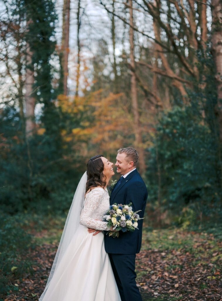 A bride and groom stand in a forest setting. The bride, in a white lace gown, holds a bouquet and smiles up at the groom, who is in a dark suit. Captured by The Woodman Inn wedding photographer, they are surrounded by tall trees with sparse autumn leaves on the ground.